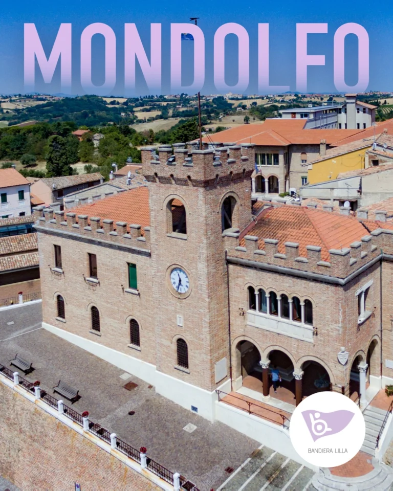 Veduta aerea del palazzo comunale fortificato di Mondolfo, in mattoncini chiari con torre dell'orologio e merlature. Sullo sfondo, le colline marchigiane e la scritta "MONDOLFO" in alto. Logo di Bandiera Lilla in basso a destra.