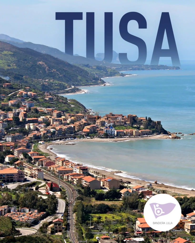 Vista panoramica dall'alto della costa di Tusa e Castel di Tusa con il mare, il borgo e le colline. In alto c'è la scritta "TUSA" e in basso a destra è presente il logo di Bandiera Lilla.