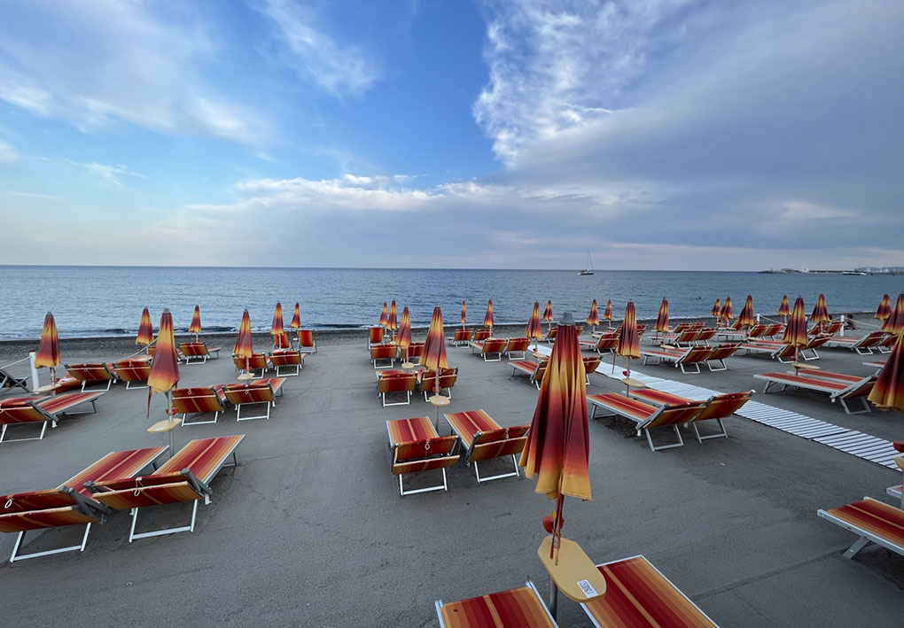 Spiaggia con file ordinate di lettini e ombrelloni nei toni del rosso, arancione e giallo; mare calmo e una barca a vela visibile in lontananza sotto cielo parzialmente nuvoloso.