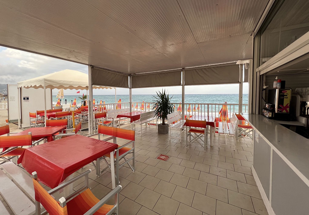 Area esterna coperta con tavoli e sedie rosse e arancioni; sullo sfondo la spiaggia con ombrelloni colorati, lettini e vista del mare.