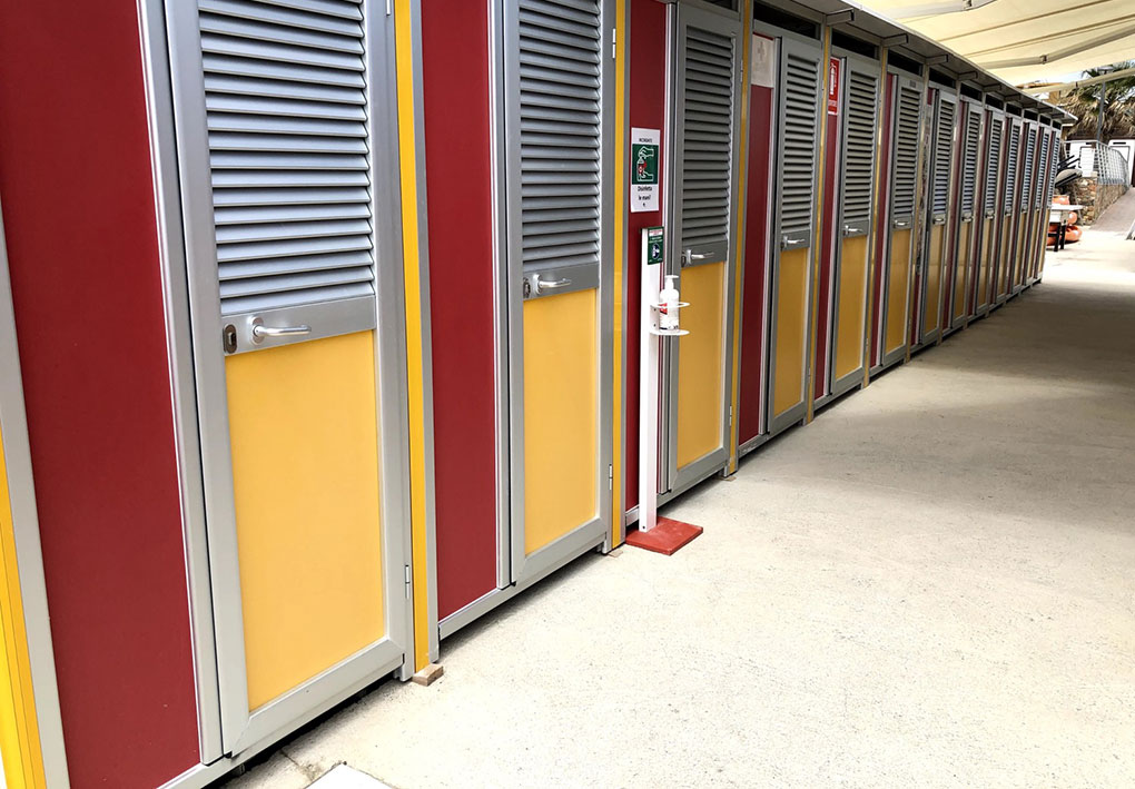 Fila di porte colorate rosse e gialle con struttura metallica, posizionate lungo un corridoio coperto con pavimento in cemento; accanto una colonnina con dispenser e segnaletica di emergenza.