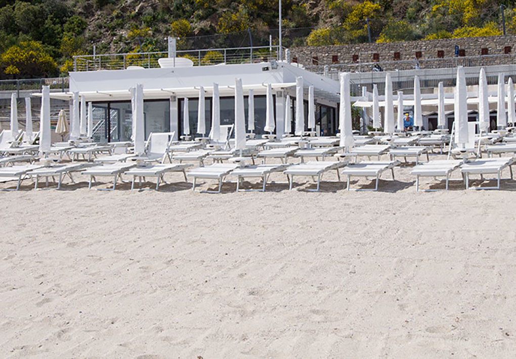 Spiaggia con file ordinate di lettini e ombrelloni bianchi davanti a una struttura moderna con ampie vetrate; alle spalle dell’edificio una collina con vegetazione e fioriture gialle.