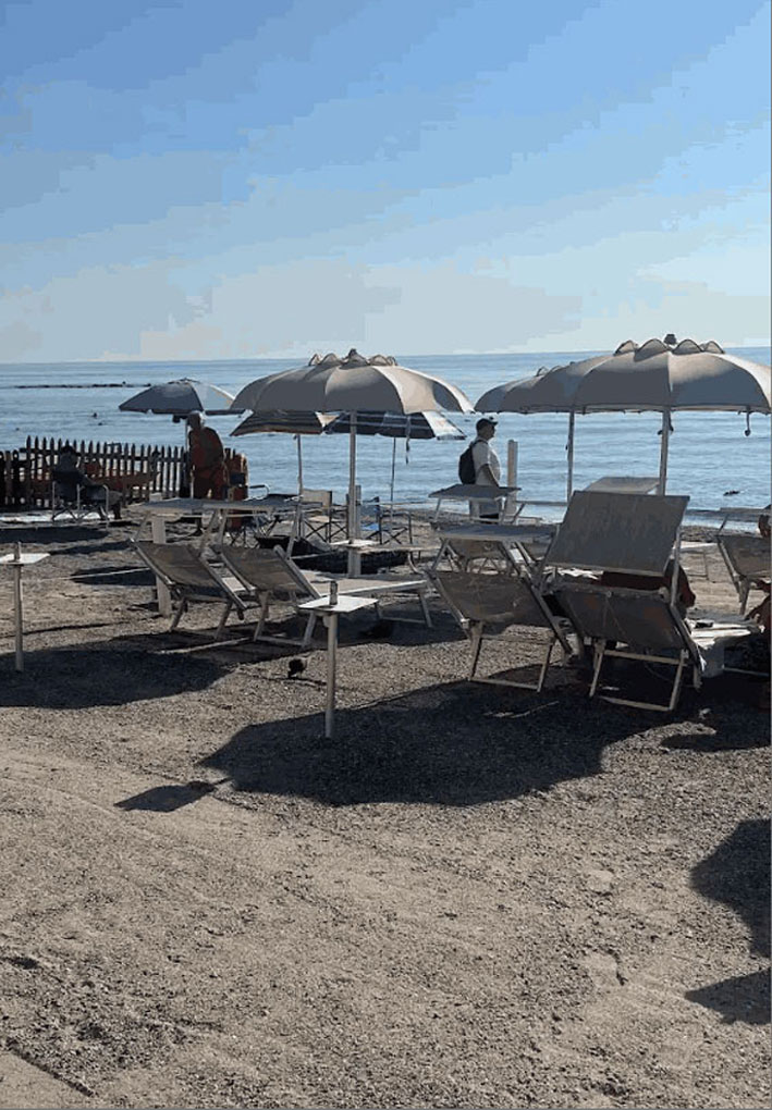 Spiaggia con lettini e grandi ombrelloni chiari disposti sulla sabbia, mare calmo e cielo limpido, alcune persone vicino alla riva e un piccolo recinto in legno sul lato sinistro.