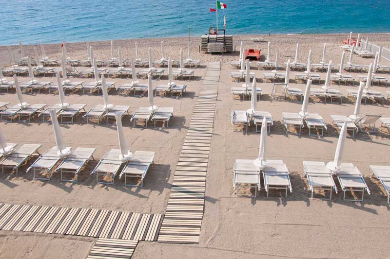 Vista frontale di uno stabilimento balneare accessibile con numerosi lettini e ombrelloni bianchi disposti in file ordinate. Una passerella in legno attraversa la spiaggia e conduce fino al mare, facilitando l'accesso alla battigia. Sullo sfondo si vede il mare calmo e una torretta di salvataggio con la bandiera italiana.