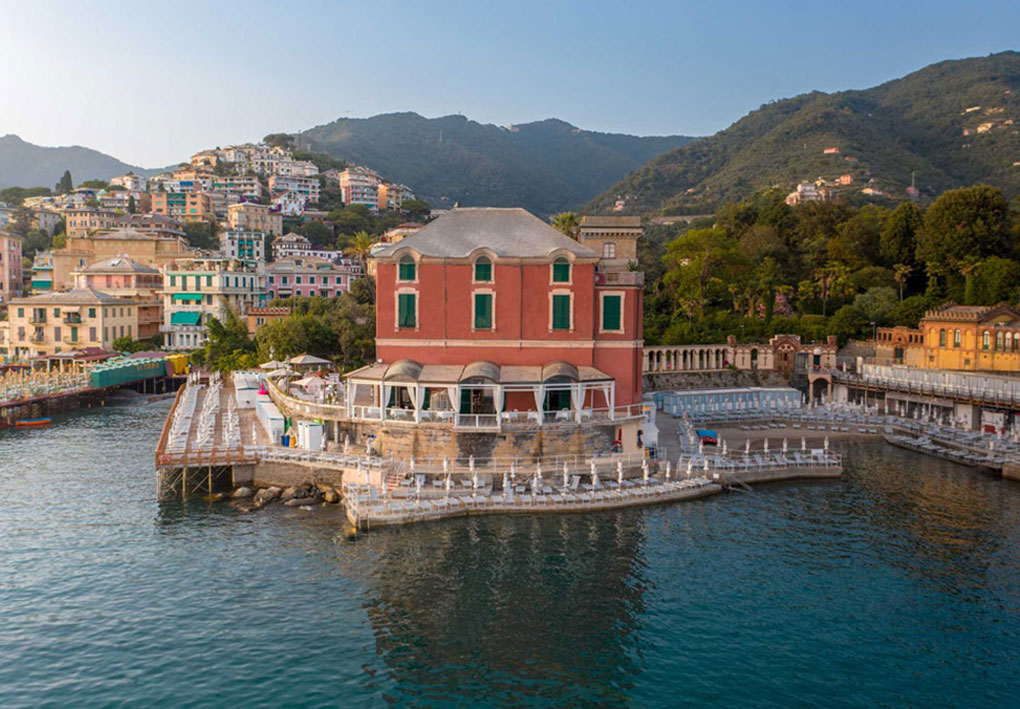 Edificio rosso affacciato sul mare, con terrazze piene di ombrelloni bianchi e lettini; alle spalle si vede il borgo colorato arrampicato sulla collina.