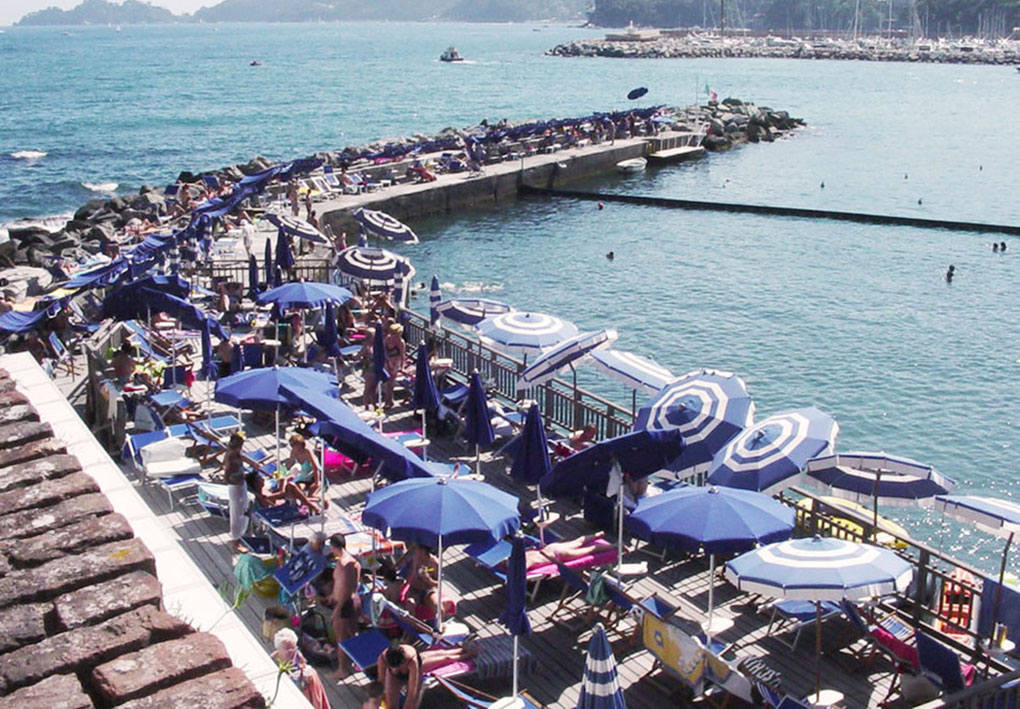 Pontile in legno affollato con ombrelloni a righe blu e bianche, persone sdraiate e bagnanti in mare; sullo sfondo la baia con barche e costa collinare.