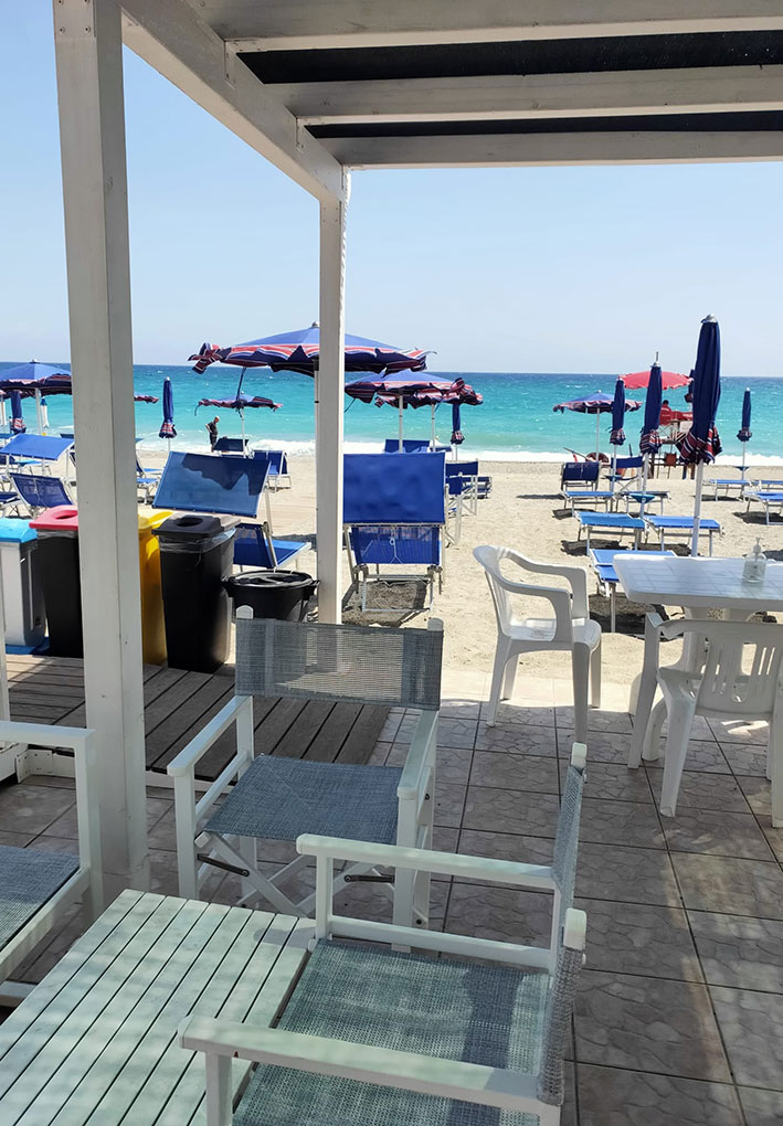 Area ombreggiata con tavoli e sedie azzurre e bianche, vista su spiaggia con lettini blu e ombrelloni viola rivolti verso mare turchese sotto cielo sereno.