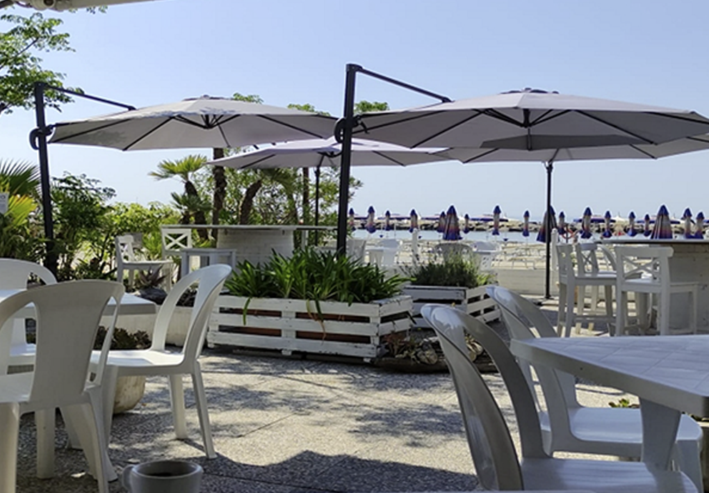 Area esterna di un bar sul mare con tavoli e sedie bianchi sotto grandi ombrelloni, affacciata su una spiaggia attrezzata con file di ombrelloni e lettini.