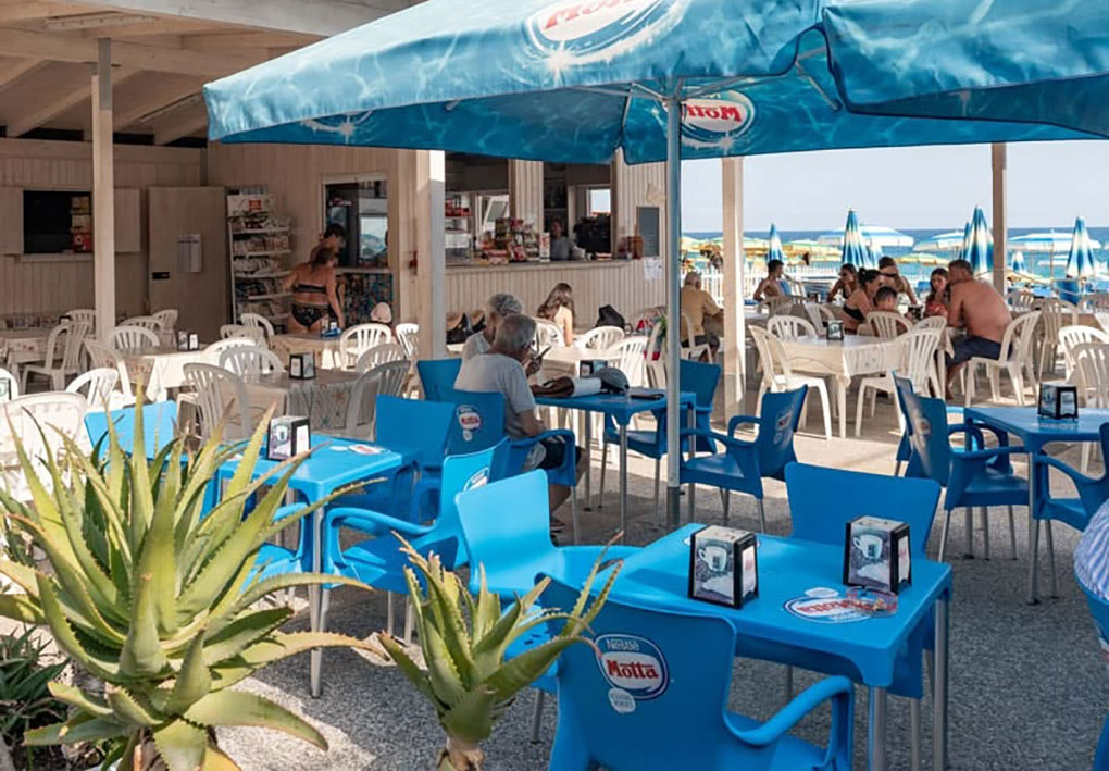 Immagine di un’area ristoro all’aperto con tavoli e sedie in plastica sotto grandi ombrelloni blu. Persone sedute consumano bevande e sullo sfondo si vedono ombrelloni e lettini in spiaggia.