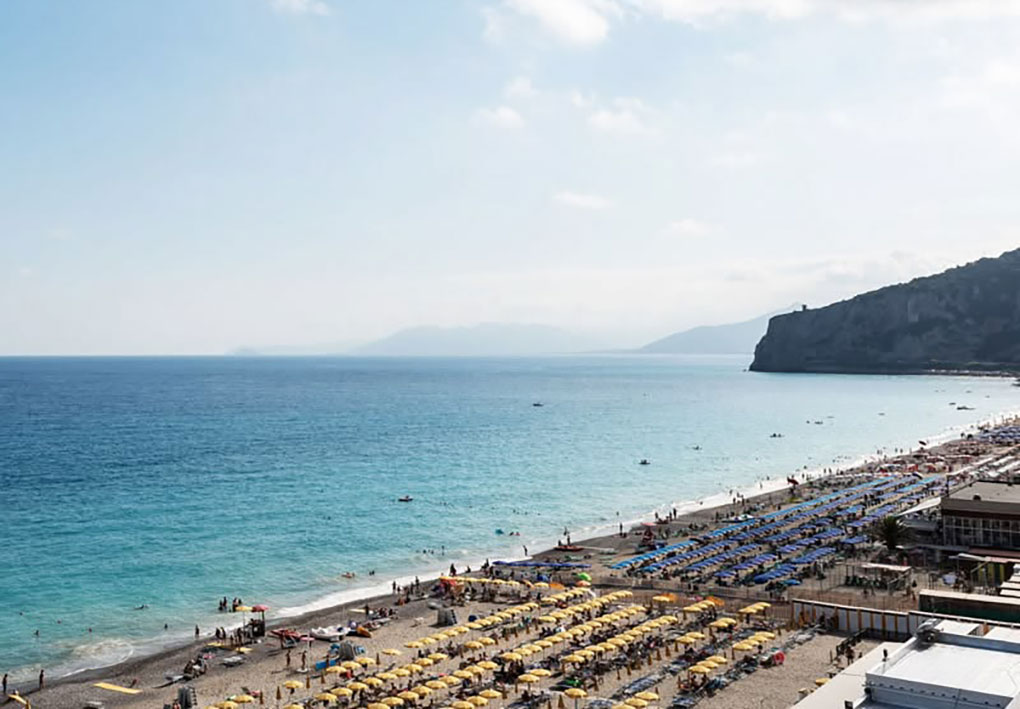 Immagine di una spiaggia affollata con file ordinate di ombrelloni colorati e persone che prendono il sole o nuotano. Sullo sfondo si nota una scogliera che si estende verso il mare.