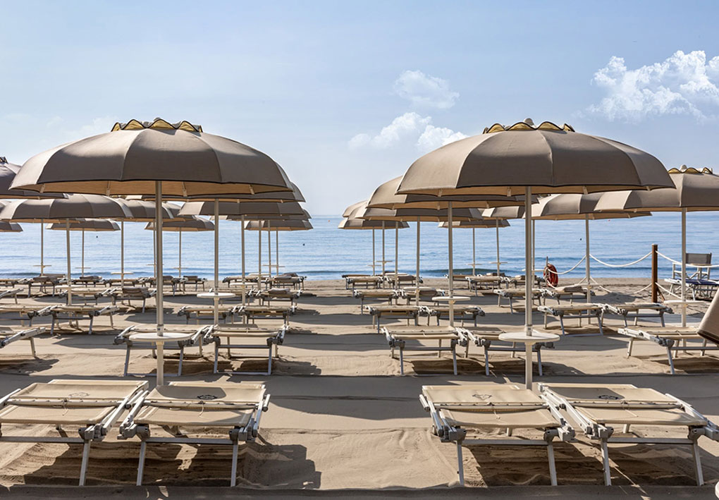 Spiaggia ordinata con file di lettini e ombrelloni beige, sabbia chiara e mare tranquillo sotto cielo sereno.