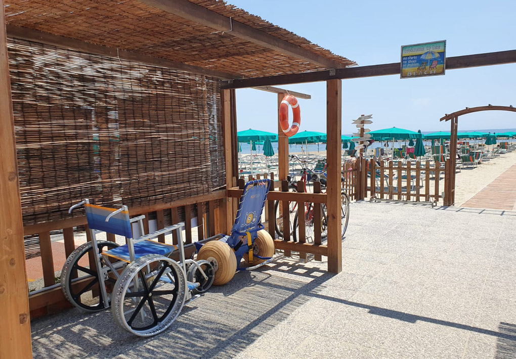 la sedia job e la sand&sea pronte per essere utilizzate all'entrata dello stabilimento, riparate da un canniccio. Oltre l'ingresso, una passerella prosegue lungo una spiaggia di sabbia con ombrelloni. sullo sfondo il mare in una giornata di sole
