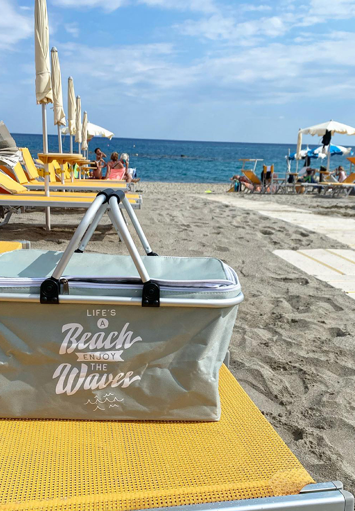 Borsa frigo grigia appoggiata su un lettino giallo, con ombrelloni e lettini gialli disposti sulla spiaggia e il mare sullo sfondo.