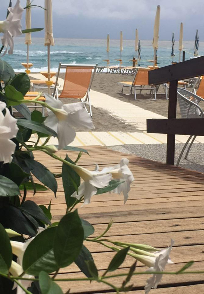 dietro una pianta fiorita decorativa, una ampia rampa di legno consente l'accesso alla spiaggia, dove file ordinate di ombrelloni sono a disposizione dei clienti. Sullo sfondo un mare con onde contro un cielo nuvoloso