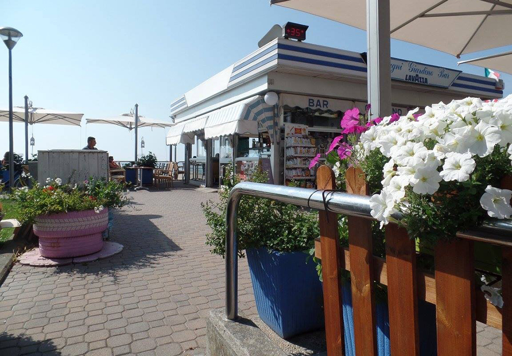 Area bar all’aperto con tende a righe bianche e blu, grandi ombrelloni, fioriere colorate e tavoli su una passeggiata pavimentata, con il mare sullo sfondo.