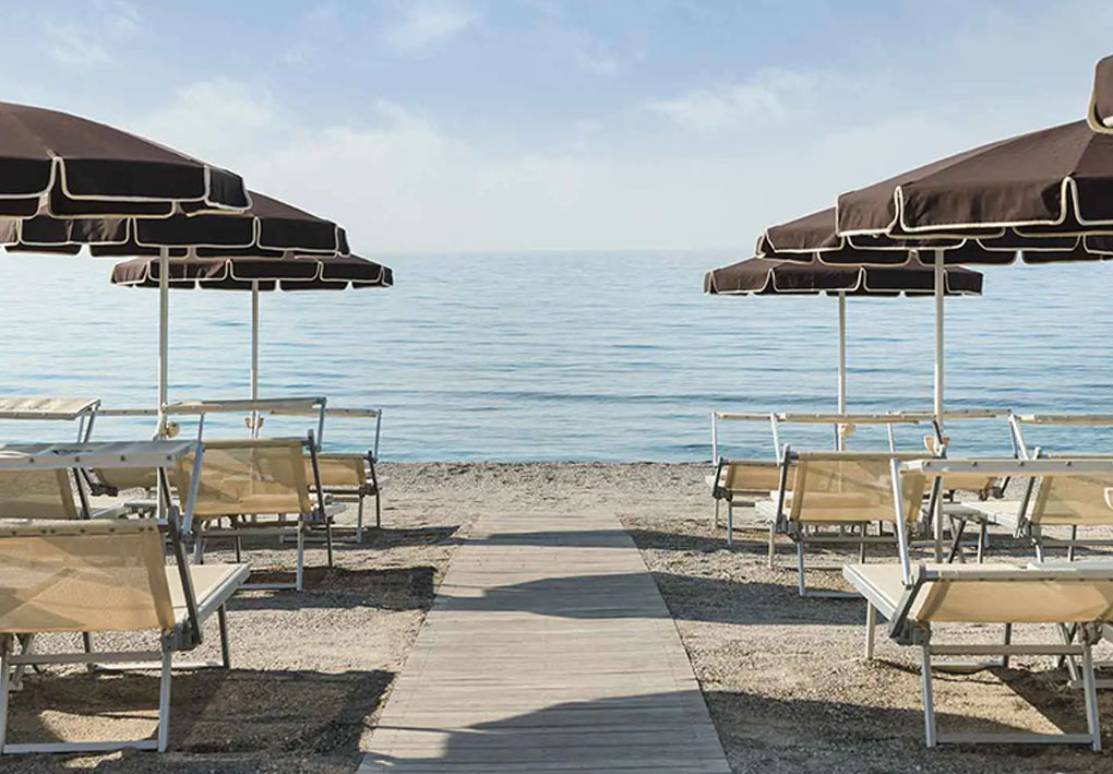 Spiaggia ordinata con file di lettini e grandi ombrelloni marroni, passerella in legno che conduce al mare calmo sotto cielo leggermente velato.