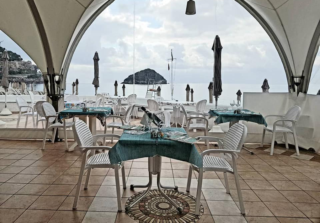 Ristorante all’aperto fronte mare con tavoli apparecchiati sotto una struttura ad archi. In fondo si vedono ombrelloni chiusi, lettini e il mare con una piccola isola sullo sfondo.