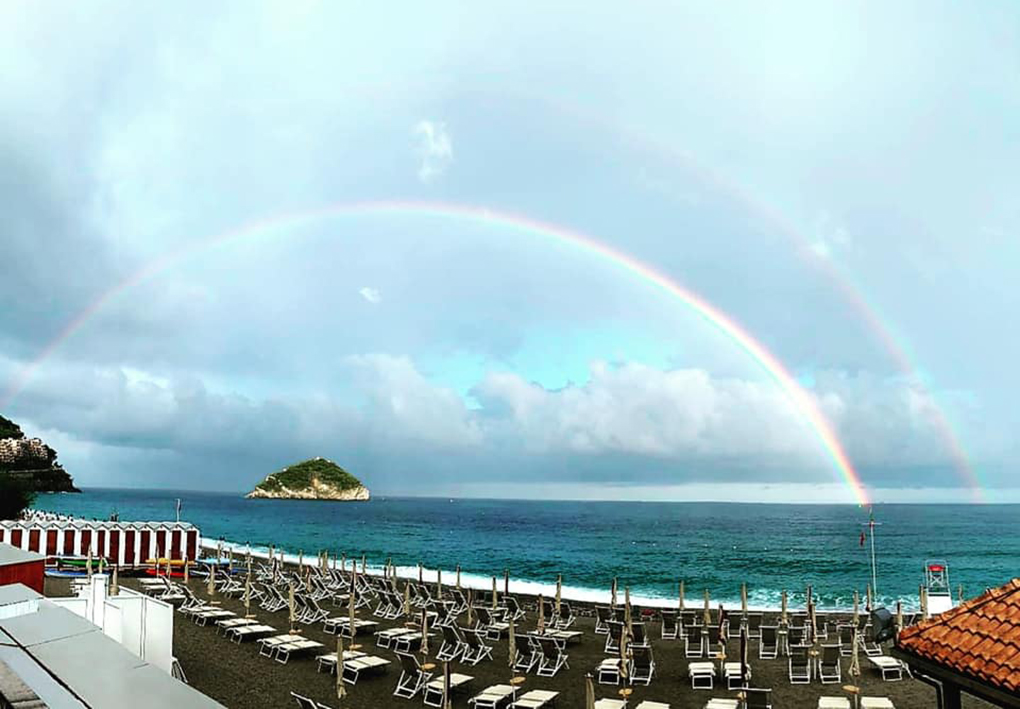 Spiaggia con file di lettini e ombrelloni rivolti verso il mare. Sullo sfondo un’isola verde e, in cielo, un doppio arcobaleno sopra l’acqua.