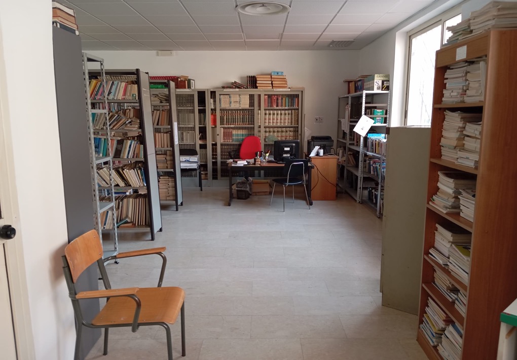 Sala della biblioteca con scaffali colmi di libri, una postazione di lavoro con computer e stampante, sedie per gli utenti e un ampio spazio libero al centro.