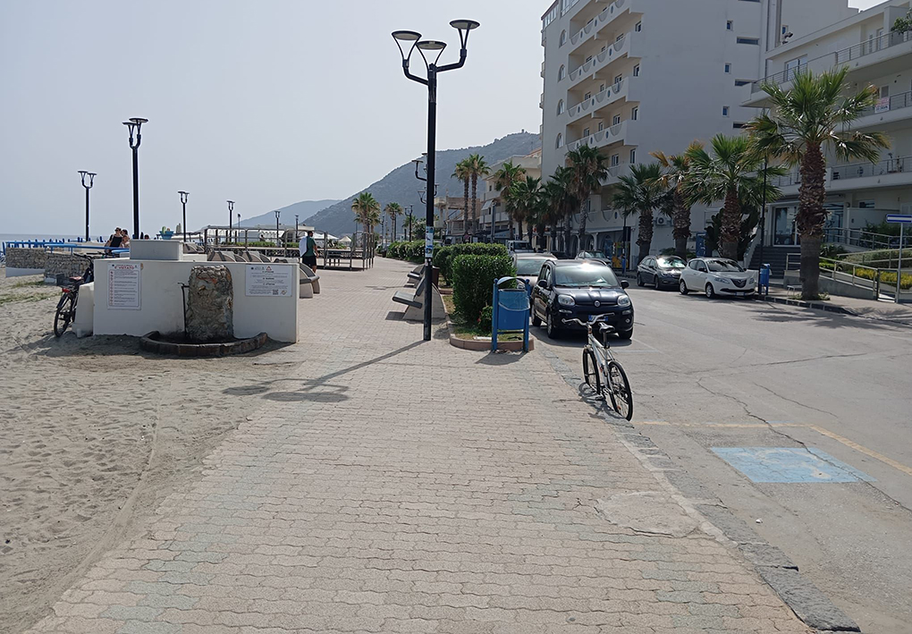 Passeggiata lungomare con pavimentazione in autobloccanti accanto alla spiaggia sabbiosa, illuminazione pubblica, palme, auto parcheggiate e edifici residenziali sul lato strada.
