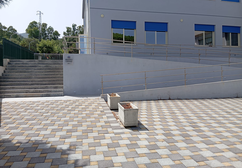 Ingresso di edificio pubblico con ampia rampa accessibile dotata di corrimano, scala laterale e piazzale pavimentato con mattonelle geometriche.