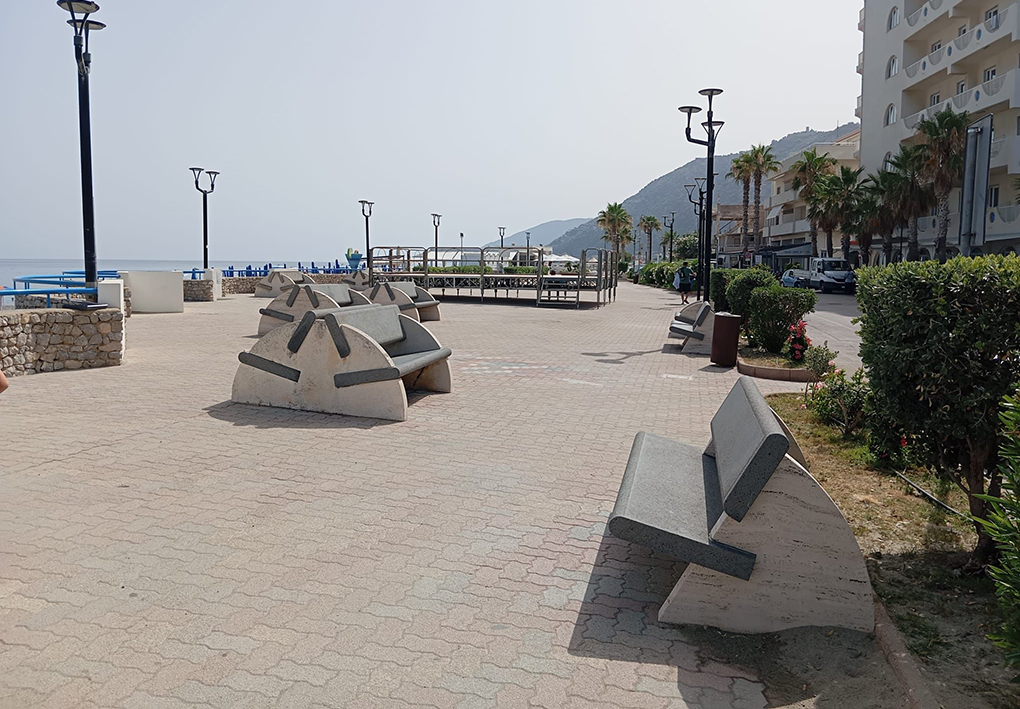 : Area del lungomare con panchine in muratura, illuminazione pubblica, aiuole e percorso pedonale pavimentato, situata tra la spiaggia e gli edifici affacciati sulla costa.