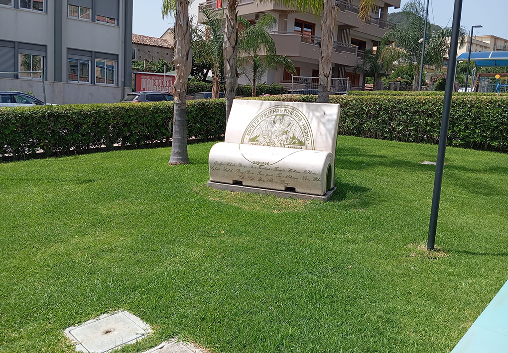 Area verde con prato e palme che ospita una panchina monumentale decorata, collocata all'interno di uno spazio urbano attrezzato e delimitato da siepi.