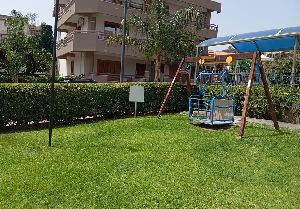 Area verde attrezzata con prato, siepi e altalena accessibile dotata di seduta con schienale, situata accanto a edifici residenziali e percorsi pedonali.
