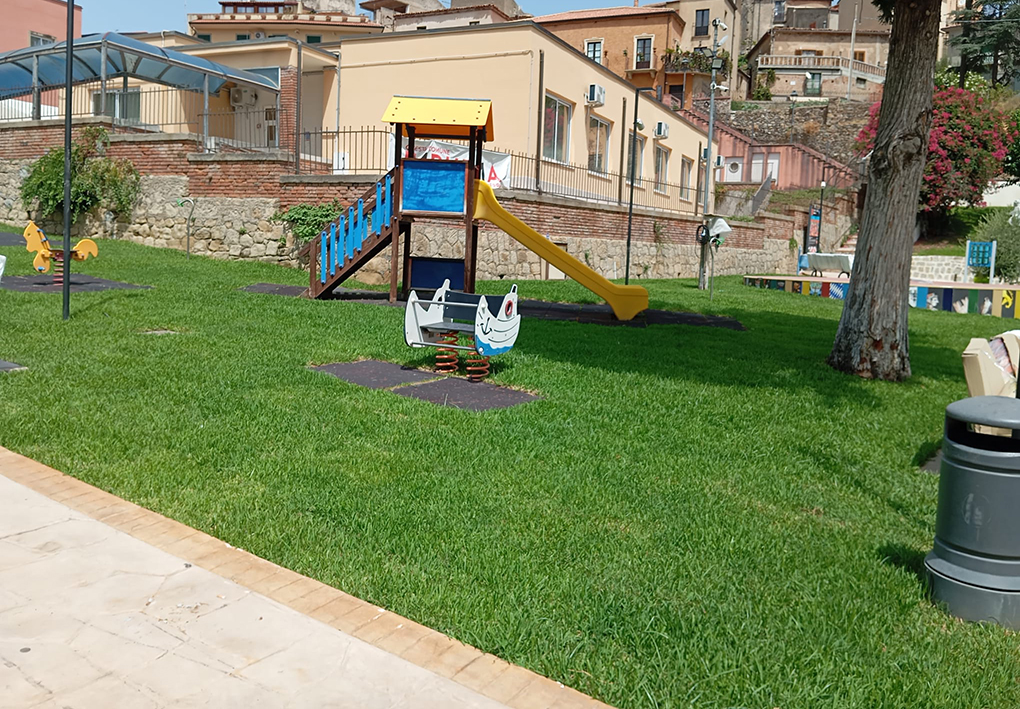Area giochi su prato con torretta dotata di scivolo e gioco a molla, inserita in uno spazio verde urbano delimitato da percorsi pavimentati.