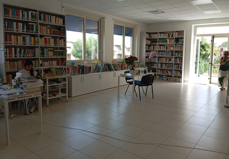 Interno di biblioteca con scaffalature colme di libri, tavolo di consultazione, ampie finestre e spazio di lettura accessibile e luminoso.