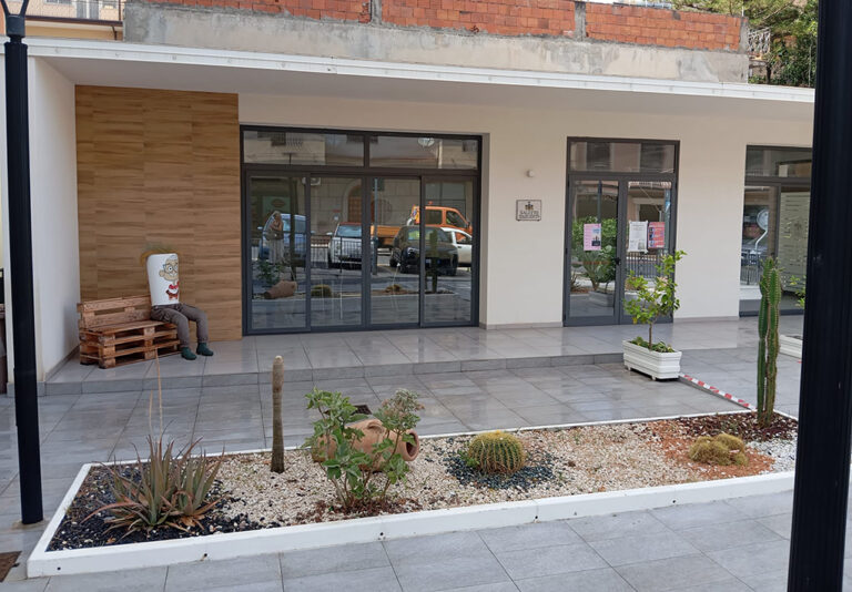 ngresso di edificio pubblico con ampia area coperta pavimentata, vetrate scorrevoli e aiuola ornamentale con piante grasse e ghiaia decorativa.