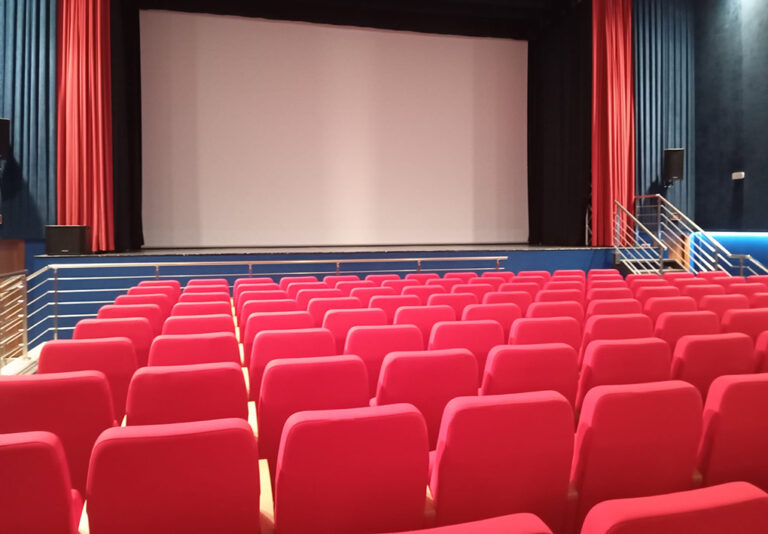 Sala teatrale con file di poltrone rosse rivolte verso il palco, dotato di grande schermo e accessi laterali con corrimano.