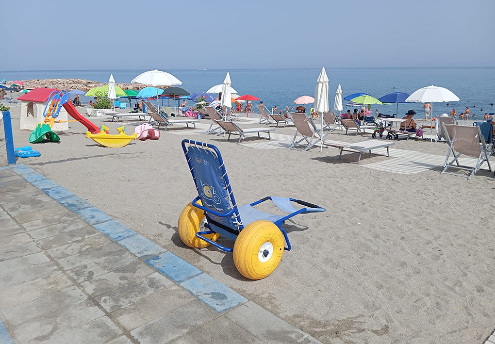 Spiaggia attrezzata con ombrelloni, lettini, giochi per bambini e sedia da mare ad anfibio posizionata vicino al percorso pavimentato.
