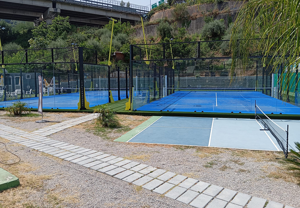 Impianto sportivo con campi da padel recintati e campo da tennis adiacente, collegati da percorsi pavimentati e circondati da vegetazione.
