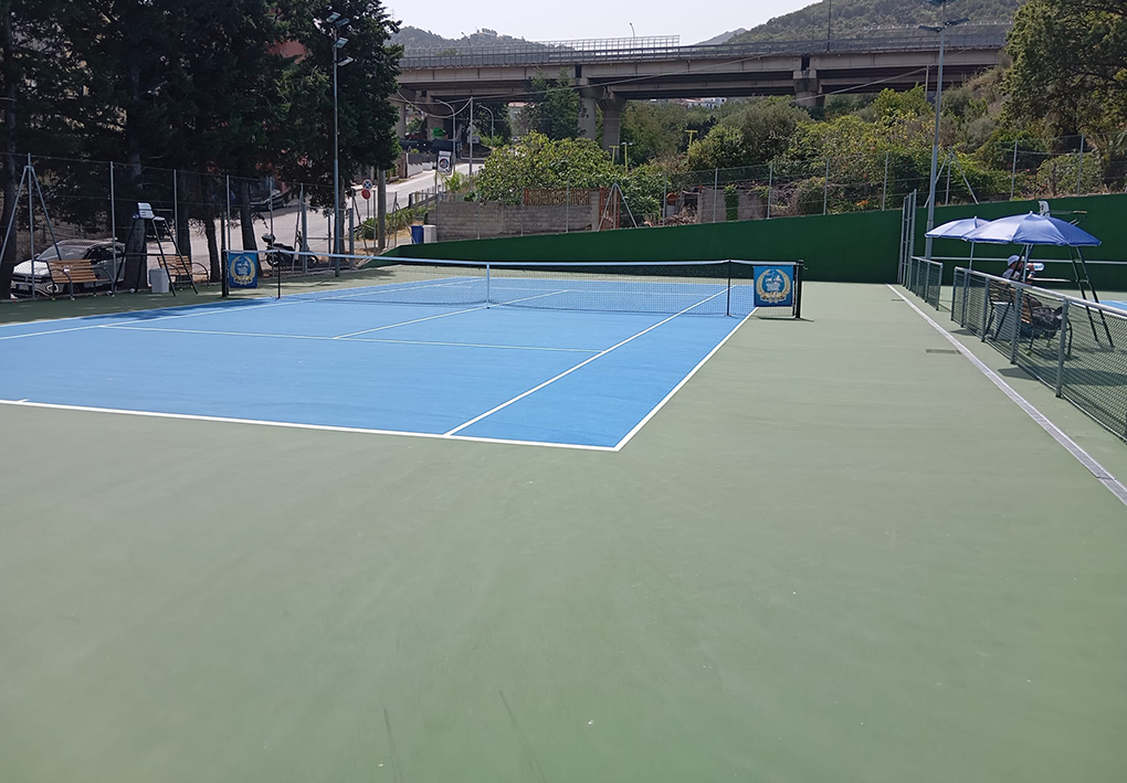 Campo da tennis all'aperto con superficie sintetica bicolore, rete centrale, recinzione perimetrale e illuminazione sportiva, inserito in un complesso sportivo circondato da vegetazione