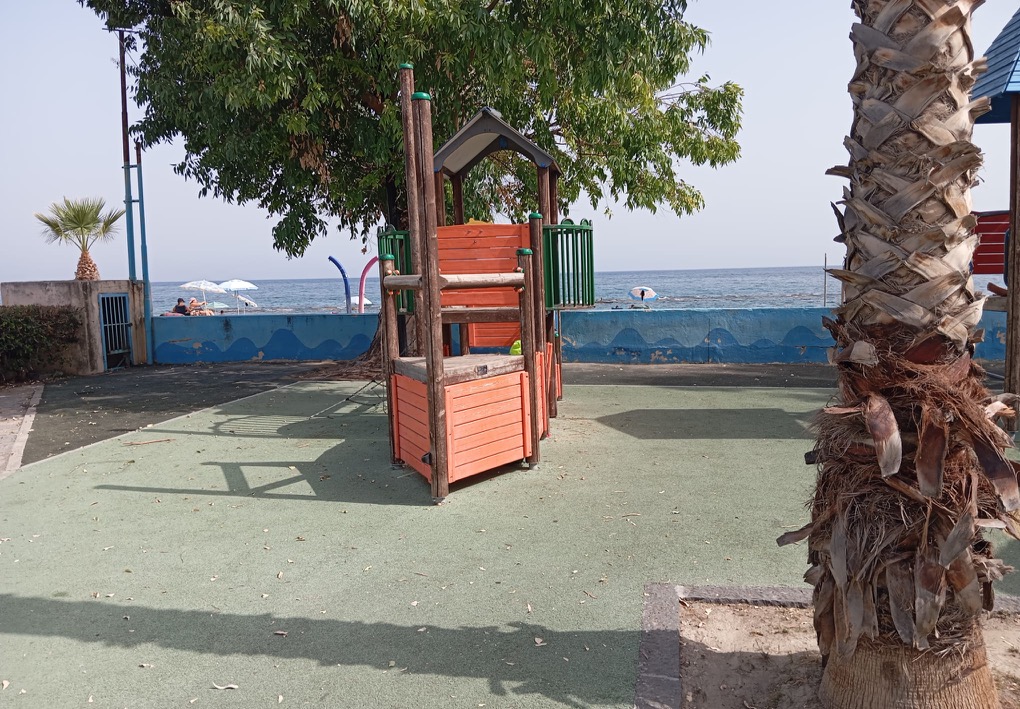 Struttura da gioco con piattaforma rialzata su pavimentazione antitrauma, circondata da alberi e palme; oltre il muretto si vedono la spiaggia, alcuni bagnanti e il mare.