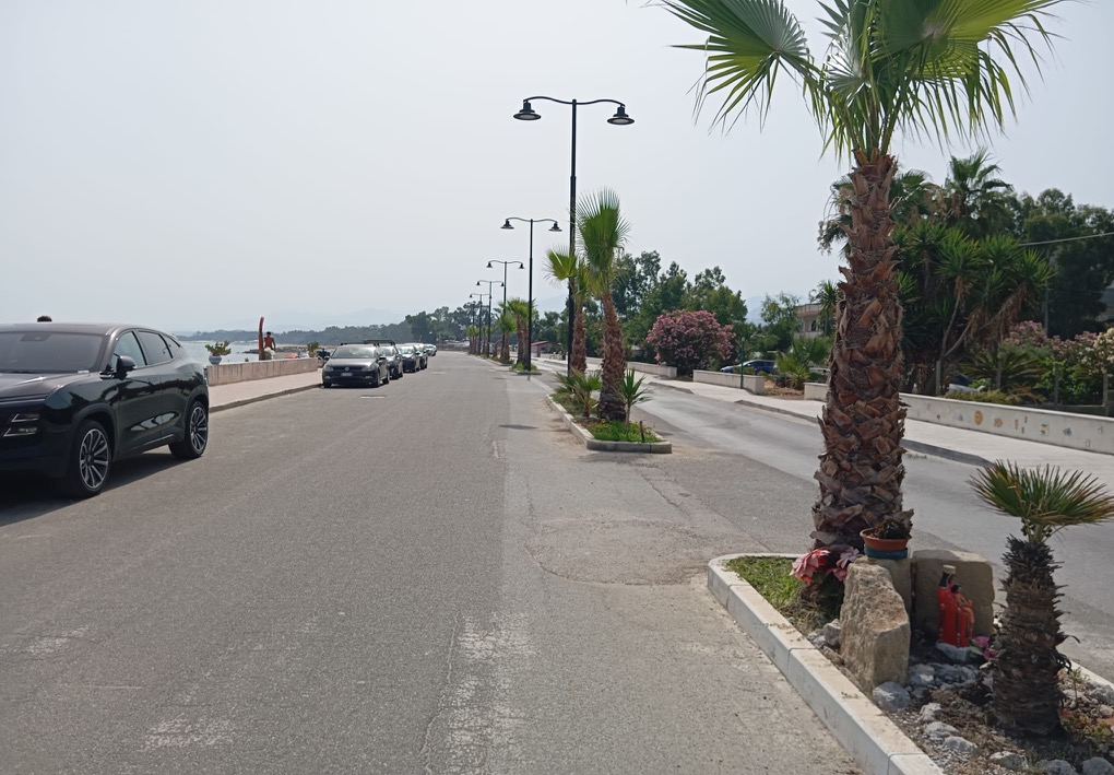 Strada sul lungomare fiancheggiata da palme e lampioni, con automobili parcheggiate lungo la carreggiata; il mare è visibile sulla sinistra, mentre sulla destra si trovano marciapiede, aiuole e giardini.