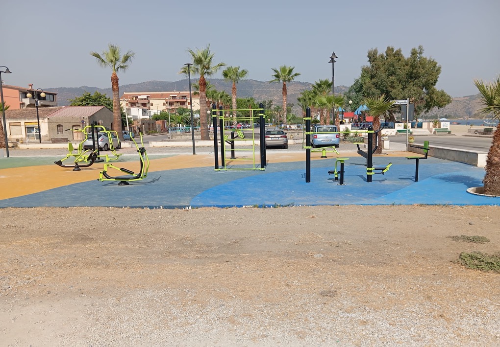Area fitness all’aperto sul lungomare, attrezzata con macchine per l’esercizio e strutture per l’allenamento; intorno si trovano palme, lampioni, automobili parcheggiate e un’area giochi, con mare e colline sullo sfondo.