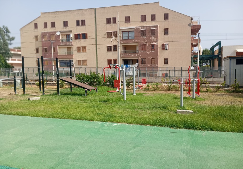 Vista panoramica di un’area fitness all’aperto con macchine per l’esercizio e strutture per l’allenamento a corpo libero, disposte su un prato.