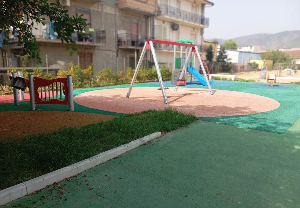Parco giochi con altalena, scivolo, gioco a molla e pannello ludico, su pavimentazione antitrauma e circondato da edifici e vegetazione.