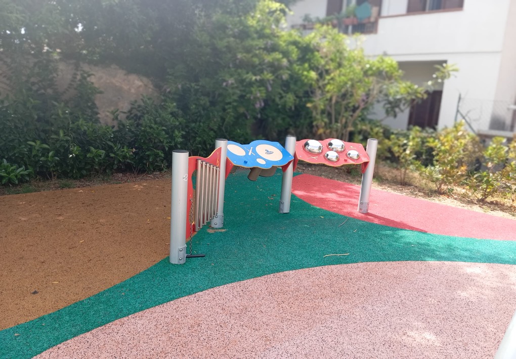 Installazioni sonore per bambini con campane, tamburi e tubi metallici, collocate su pavimentazione antitrauma accanto a siepi e alberi.