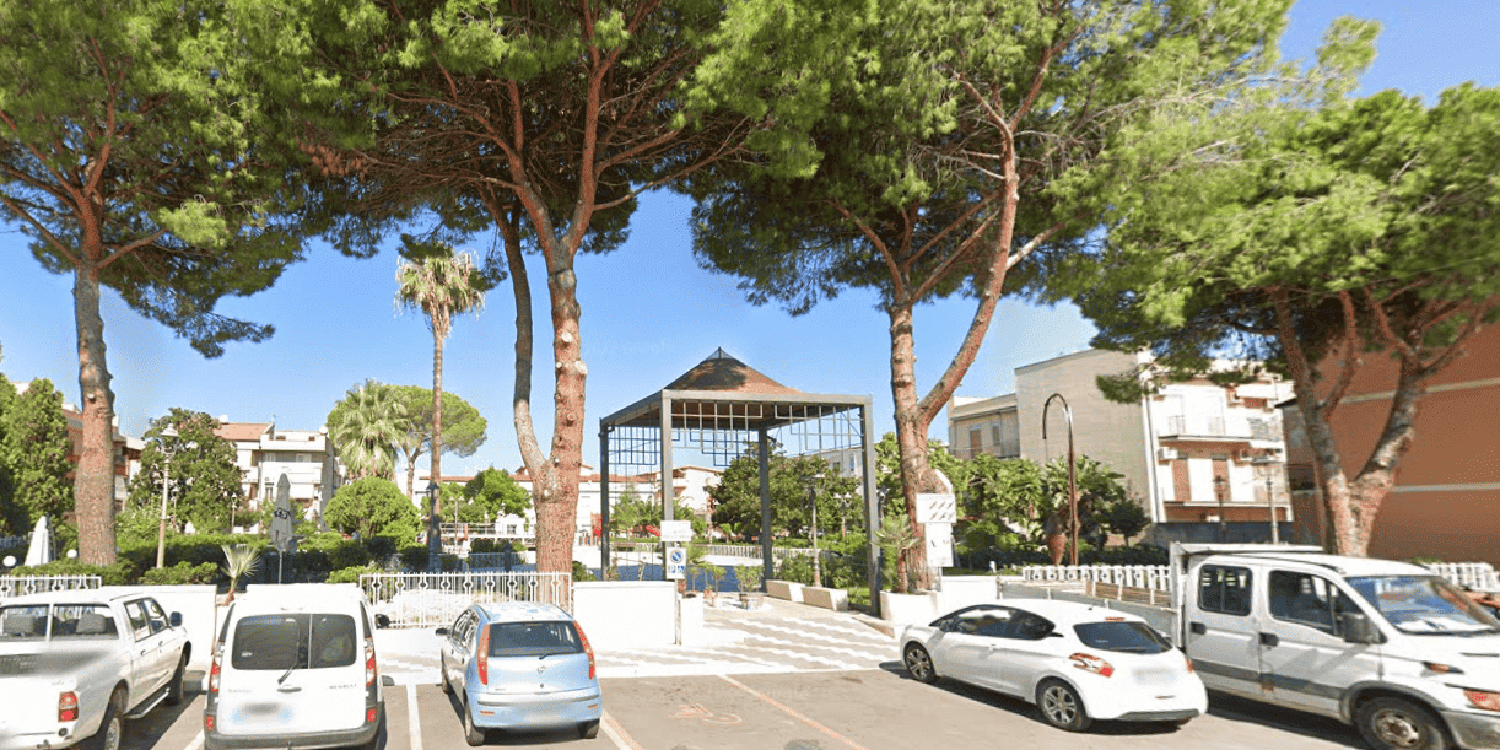 Ingresso di Piazza Principe Romeo, ombreggiata da grandi pini e palme, con un gazebo dalla struttura metallica al centro, aiuole curate, panchine, edifici circostanti e automobili parcheggiate lungo la strada.