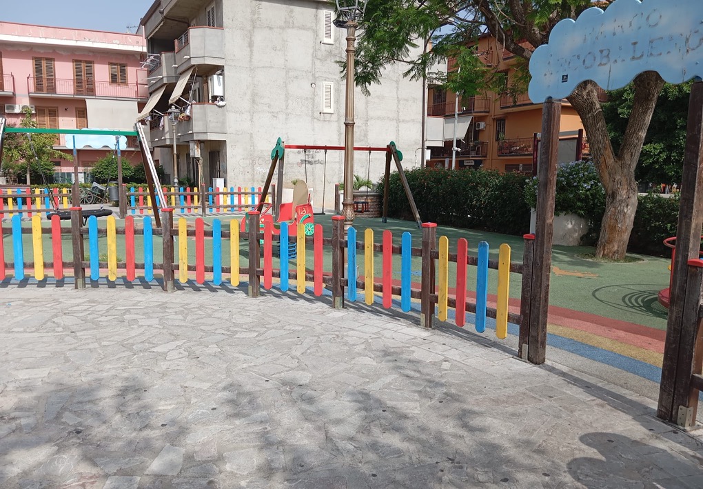 Vista esterna di un parco giochi recintato con altalene, un gioco a molla, alberi e pavimentazione antitrauma.