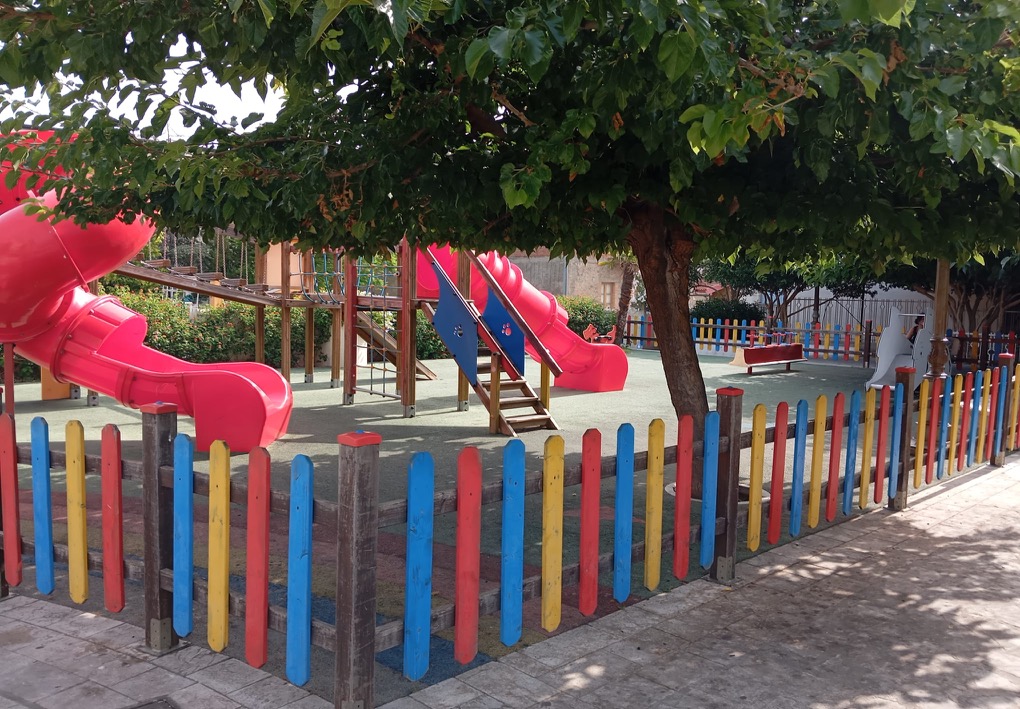Parco giochi ombreggiato da un grande albero, con scivoli, strutture per arrampicarsi e altri giochi su pavimentazione antitrauma, delimitato da una recinzione.
