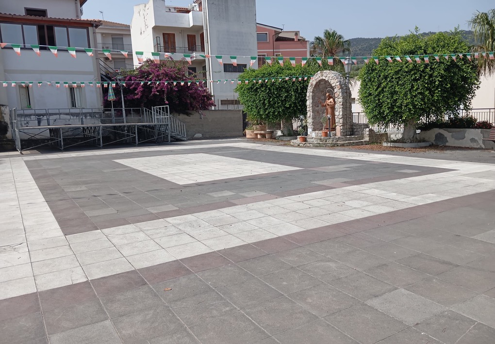 Piazza pedonale pavimentata con una statua religiosa collocata in una nicchia di pietra, alberi potati, panchine e festoni sospesi; sullo sfondo si trovano abitazioni e un palco rialzato con rampa.