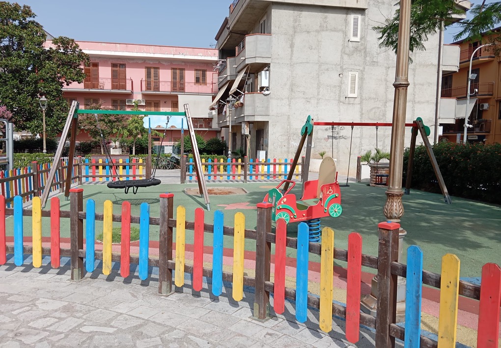 Parco giochi recintato con altalene, una seduta a cesto e un gioco a molla accessibile, su pavimentazione antitrauma e circondato da edifici residenziali.
