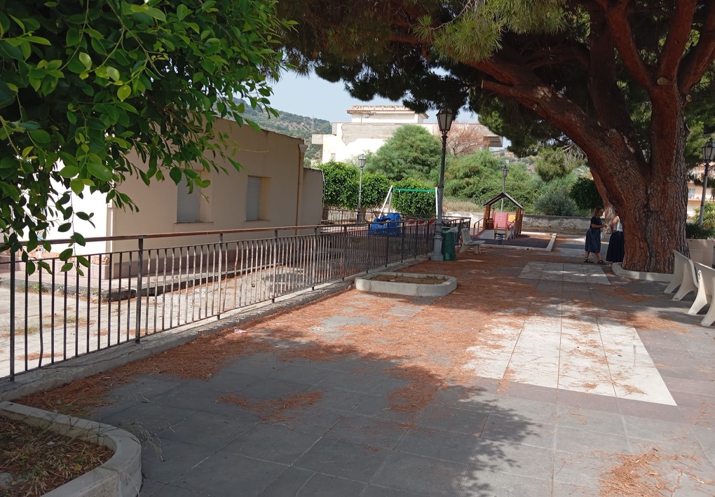 Viale pedonale ombreggiato da grandi pini, con panchine, lampioni e una ringhiera laterale; sullo sfondo si trovano un’area giochi e due persone accanto a un albero.
