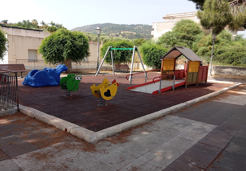 Area giochi all’aperto con pavimentazione antitrauma, giochi a molla, un’altalena e una casetta rialzata raggiungibile tramite rampa; intorno si trovano panchine, alberi ed edifici, con una collina sullo sfondo.