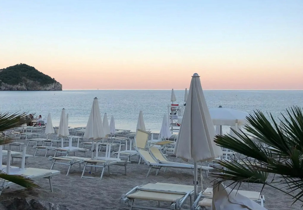 Spiaggia al tramonto con file di lettini e ombrelloni chiusi, foglie di palma in primo piano, promontorio roccioso sulla sinistra e mare calmo che si estende fino all'orizzonte.