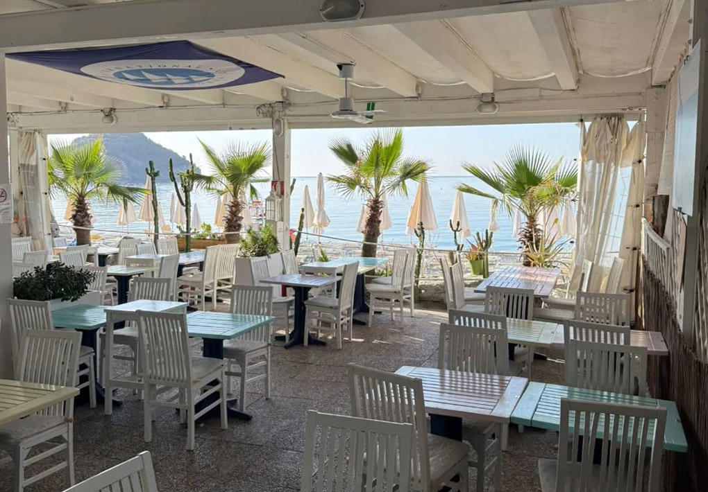 Terrazza coperta di un locale sul mare, con tavolini e sedie disposti sul pavimento in pietra, palme e piante in vaso lungo il perimetro, con apertura sulla spiaggia e sul mare, di fronte all'isola di bergeggi